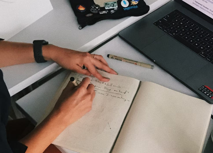Person writing in notebook beside laptop
