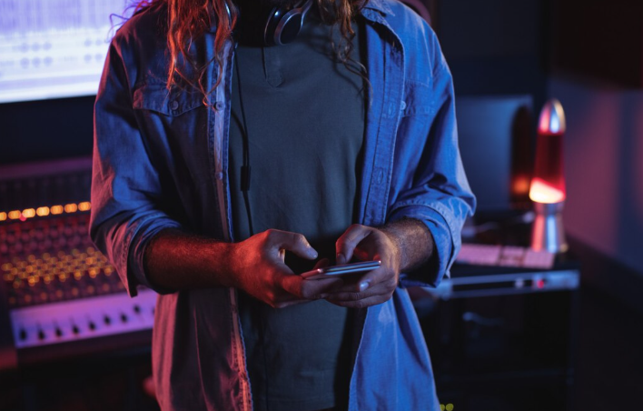 Man in recording studio using smartphone near audio equipment.