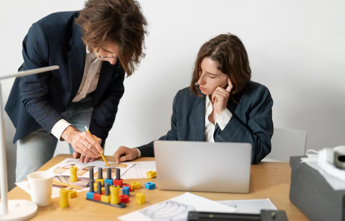 Two colleagues discussing project details with documents and laptop on desk.