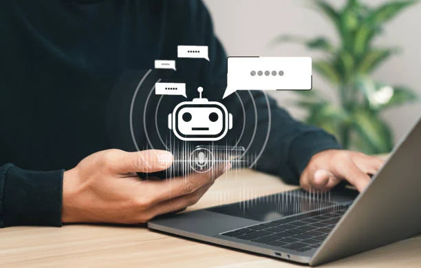 Person using smartphone with chatbot icon and laptop on wooden desk.