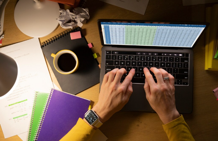 Person working on laptop with spreadsheets, notebooks, and coffee on desk.