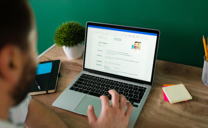 Man editing resume document on laptop at desk with notepad.