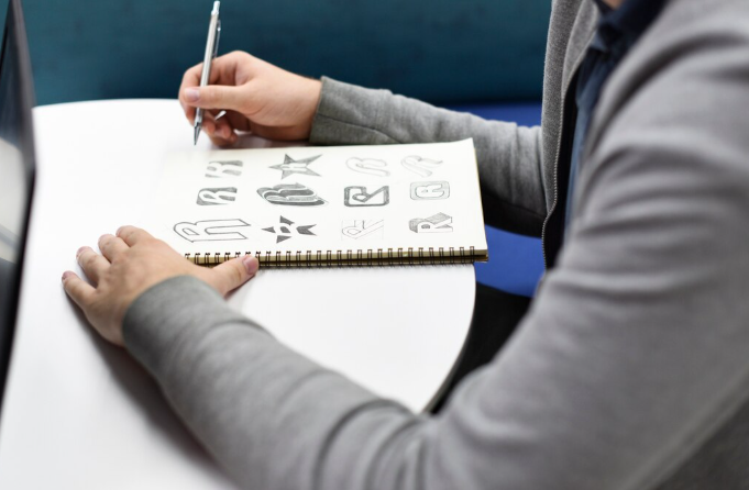 Designer sketching logo concepts in a notebook with different letter and icon variations.
