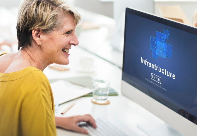 Smiling woman working at a desktop computer displaying the word “Infrastructure” on screen.