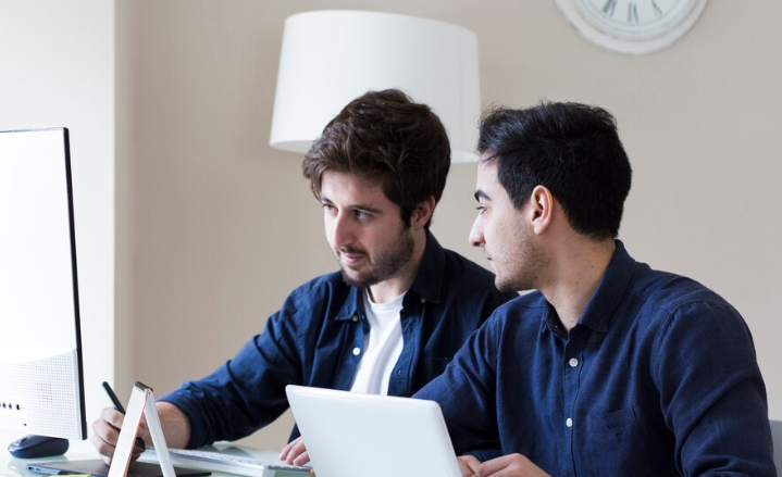 Two young men working together at a desk with a desktop computer and a laptop, collaborating on a project.