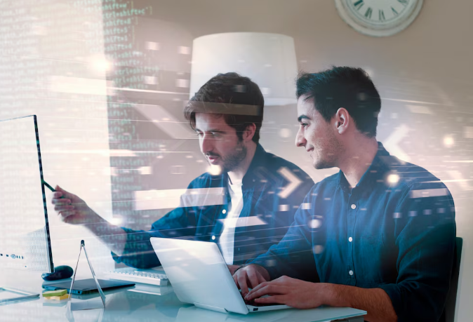 Two men collaborating in front of monitors with a transparent overlay of programming code.