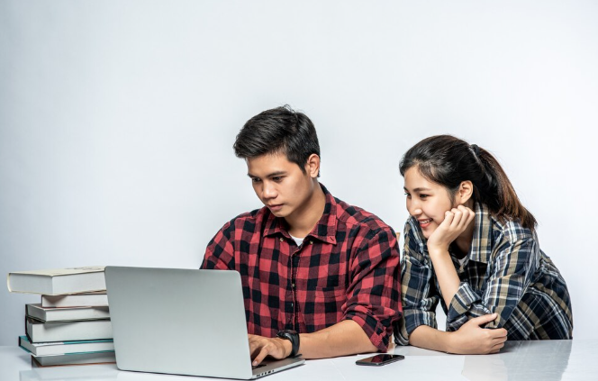 Two students using laptop together with books stacked nearby.