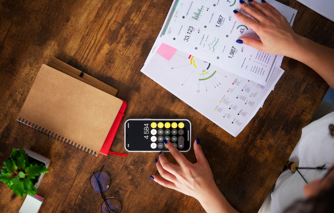 Person reviewing graphs with smartphone calculator and reports on wooden table.