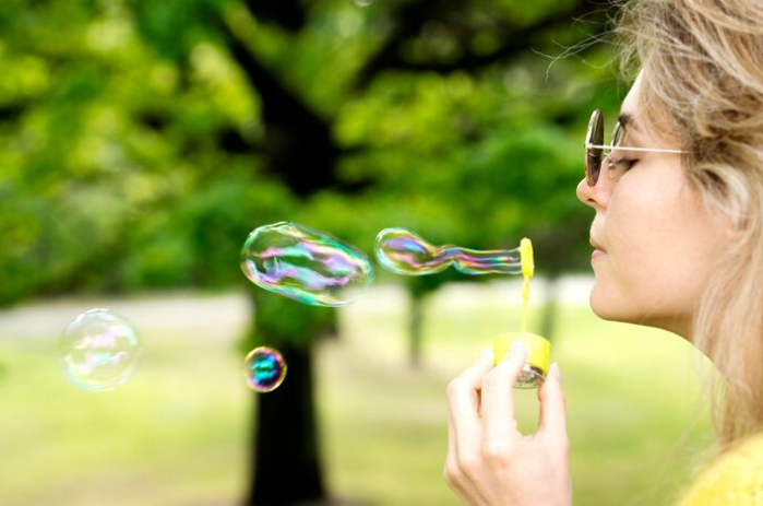 Woman wearing glasses blowing colorful soap bubbles outdoors in park.