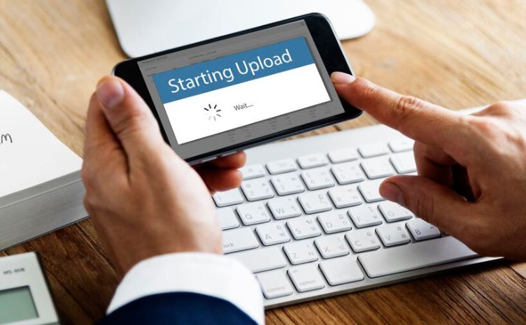 Person using smartphone to start file upload beside computer keyboard.