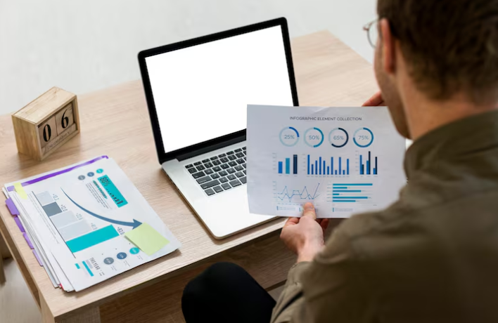 Man reviewing printed infographic charts while working on laptop.
